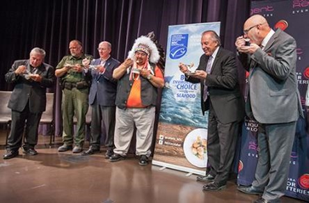 Six dignitaires, dont un chef autochtone, aux côtés d’une enseigne sur une scène. Chaque personne goûte à du brochet dans une petite assiette.