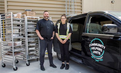 Two people stand beside a black delivery van labeled “Community Helpers Unite – Feeding Community” at a loading dock.
