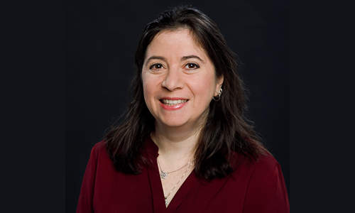 A woman in front of a dark background is wearing a burgundy blouse, two gold necklaces, and hoop earrings.  She is smiling at the camera.