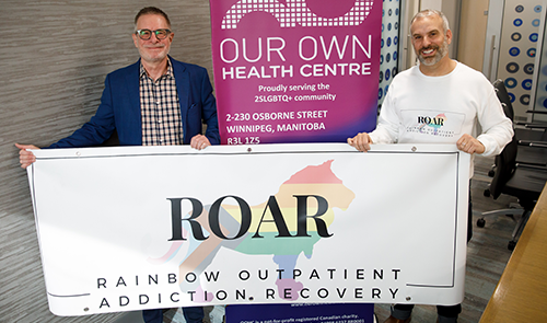 Two smiling individuals stand indoors holding a banner that reads "ROAR - Rainbow Outpatient Addiction Recovery."